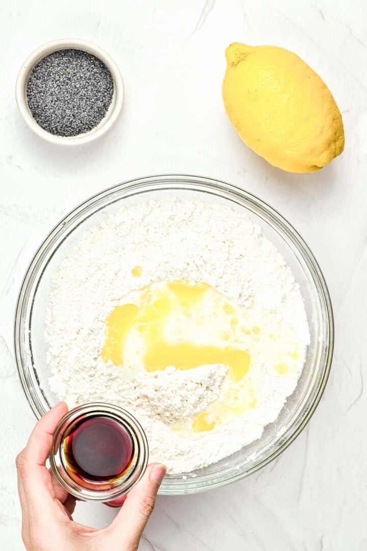 Top view of flour mixture in a bowl, a hand pouring vanilla, with a lemon and poppy seeds nearby on a white surface.
