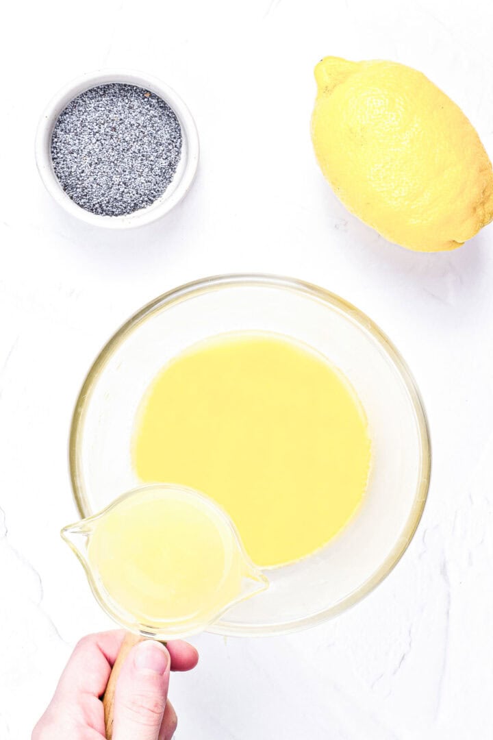 Hand pouring lemon juice into a bowl with lemon mixture; lemon and poppy seeds nearby on a white surface.