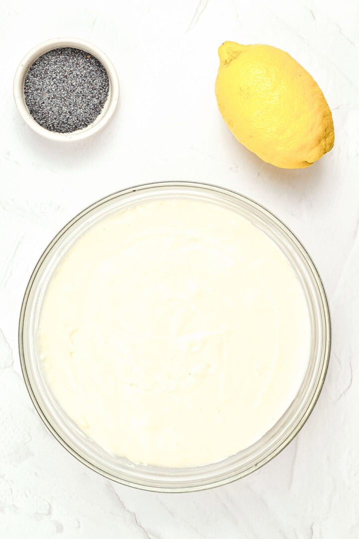 A bowl of creamy batter, a lemon, and a small bowl of poppy seeds on a white surface.