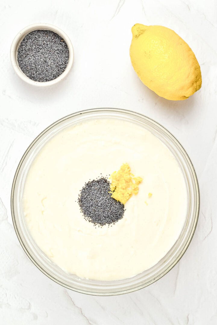 A bowl of creamy batter with poppy seeds and lemon zest, next to a whole lemon and a small bowl of poppy seeds.