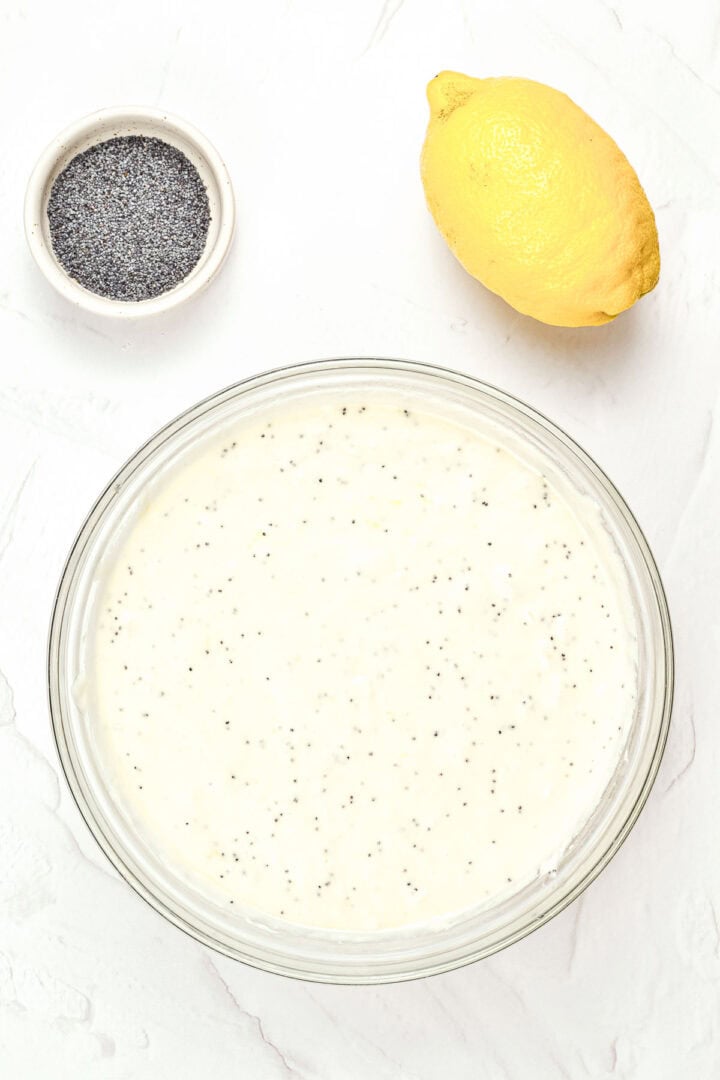 A bowl of lemon poppy seed batter, a whole lemon, and a small bowl of poppy seeds on a white surface.