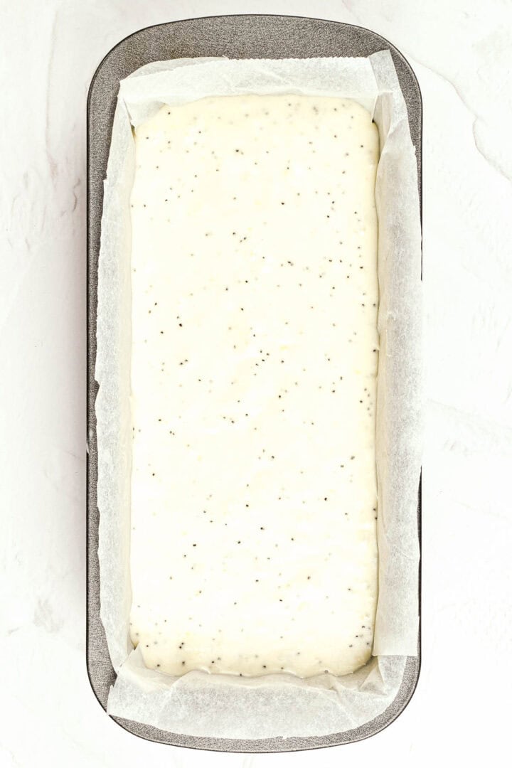 Rectangular loaf pan lined with parchment paper, filled with speckled white batter ready for baking.