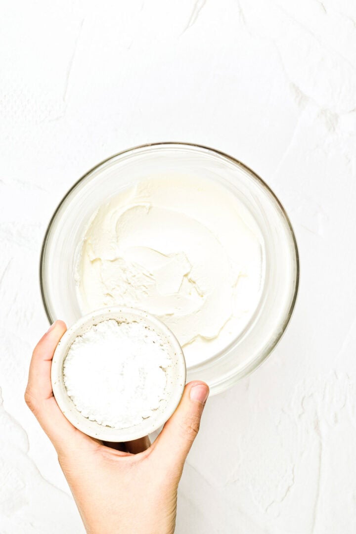 A hand holds a small bowl of powdered sugar above a glass bowl of whipped cream on a white surface.