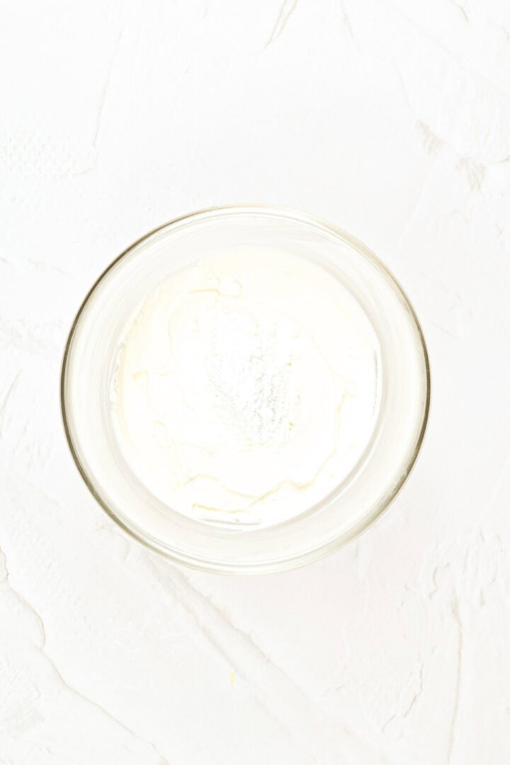 A glass bowl filled with smooth white cream on a textured white surface, viewed from above.