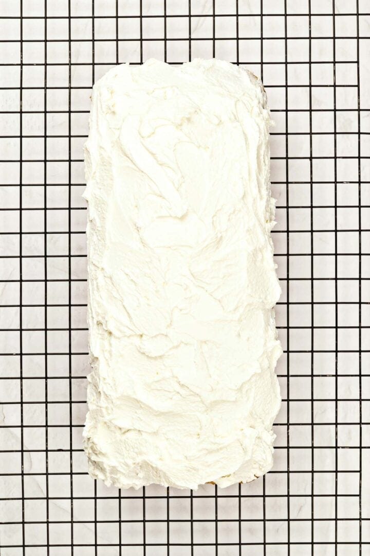 Rectangular loaf covered in white frosting, cooling on a black wire rack over a white surface.