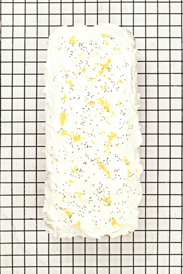 Rectangular loaf cake with white frosting, lemon zest, and poppy seeds on a black wire cooling rack.