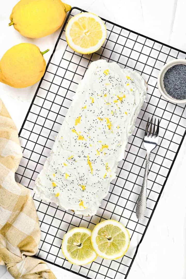 Lemon poppy seed loaf with white icing on a cooling rack, surrounded by lemons and a fork.