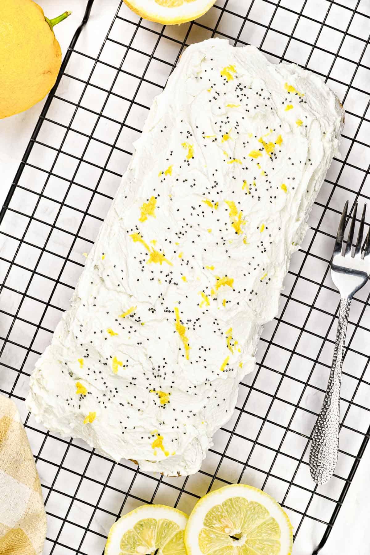 Rectangular lemon poppy seed cake with frosting, lemon zest, and poppy seeds on a cooling rack, with lemons nearby.