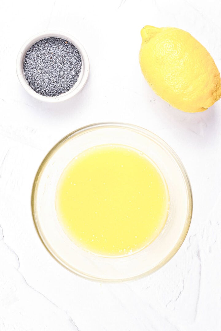A bowl of lemon juice, a whole lemon, and a small dish of poppy seeds on a white surface.