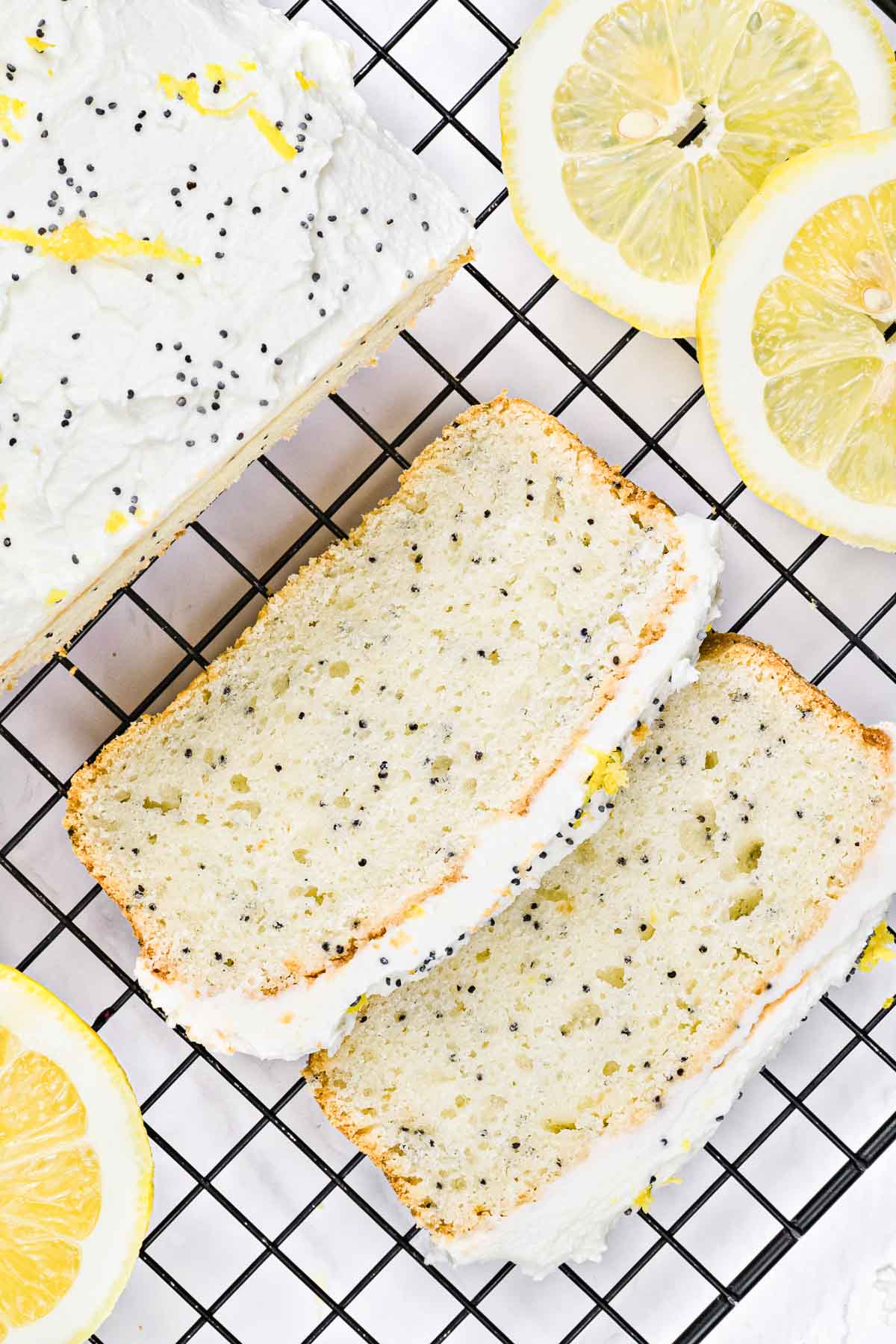 Two slices of lemon poppy seed cake with icing on a cooling rack, next to fresh lemon slices.