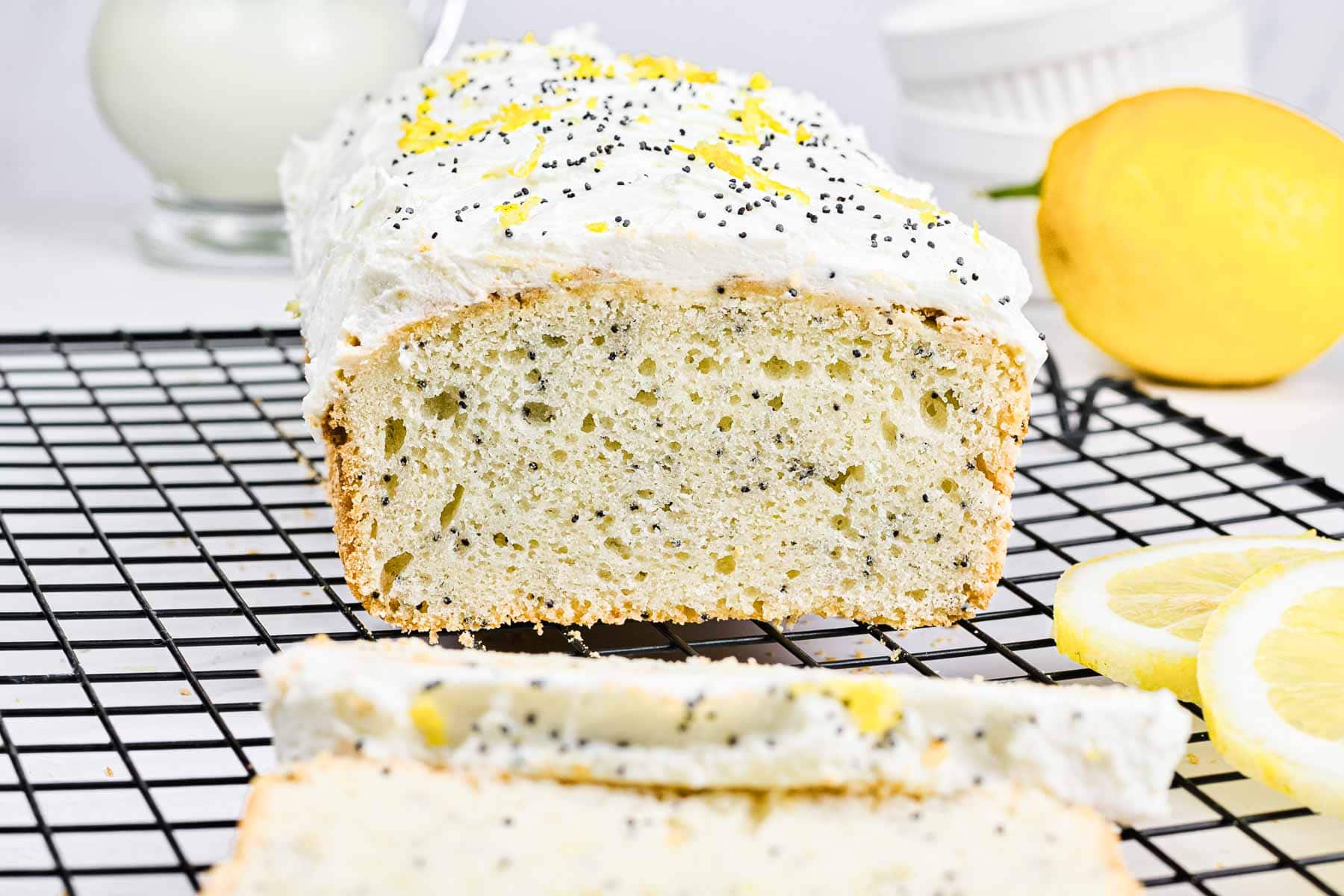 A loaf of lemon poppy seed bread with icing, sliced open, on a cooling rack next to lemon slices.