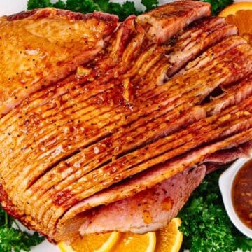 Sliced glazed ham on a platter with orange slices, parsley, and a small bowl of sauce.