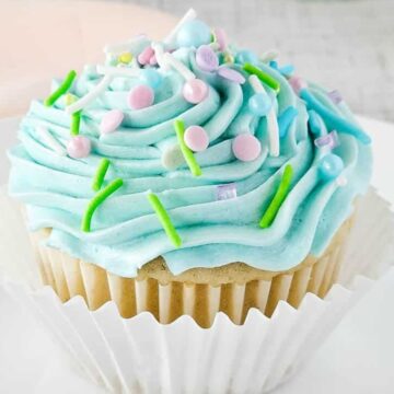 Vanilla cupcake with blue frosting and colorful sprinkles on a white plate, in a paper cupcake liner—a delightful treat perfect for Easter sweets.
