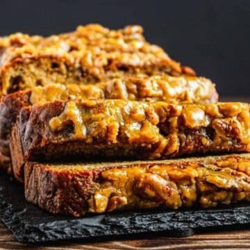 Sliced banana bread loaf topped with walnuts, displayed on a black slate board on a wooden table.