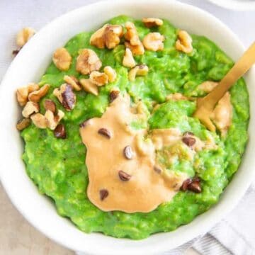 A bowl of green oatmeal topped with peanut butter, chopped walnuts, and chocolate chips, with a gold spoon.