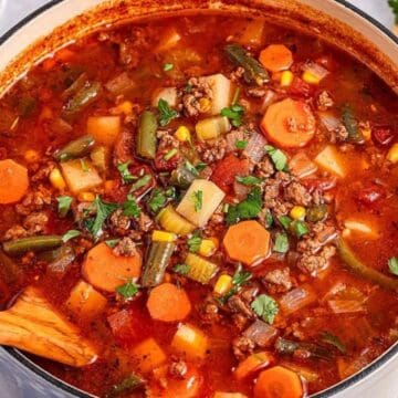 A pot of hearty vegetable beef soup with carrots, potatoes, corn, and herbs, with a wooden spoon stirring.