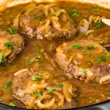 Four Salisbury steaks with gravy, onions, and parsley in a skillet with a serving spoon.