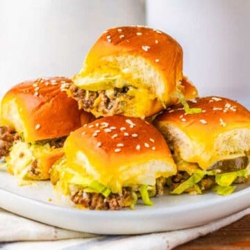 Four cheeseburger sliders with lettuce and pickles on a white plate, topped with sesame seed buns.