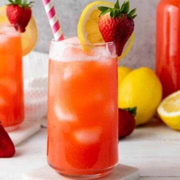 Two glasses of strawberry lemonade with ice, lemon slices, and strawberries, with striped straws on a white table.