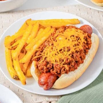 Chili cheese hot dog with French fries on a white plate, set on a beige placemat next to a green napkin.