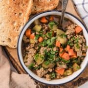 Bowl of vegetable and ground meat soup garnished with herbs, served with slices of toasted bread.