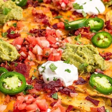 Loaded nachos topped with jalapeños, guacamole, sour cream, bacon, tomatoes, and melted cheese on a tray.