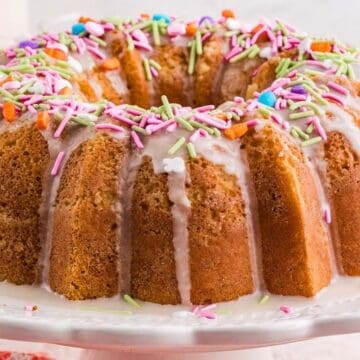 Bundt cake with white glaze and colorful sprinkles on a white cake stand.