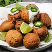 Falafel balls on a plate, topped with sauce, sliced jalapeños, cilantro, and lime wedges.