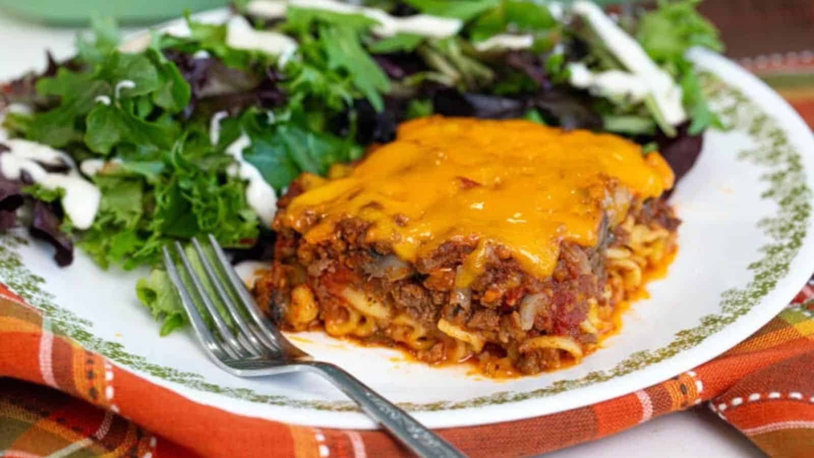 A slice of cheesy lasagna with meat sauce served next to a green salad with dressing on a white plate.