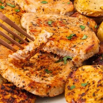 Seasoned pork chops and roasted potatoes garnished with parsley on a white plate, with a fork cutting a piece.