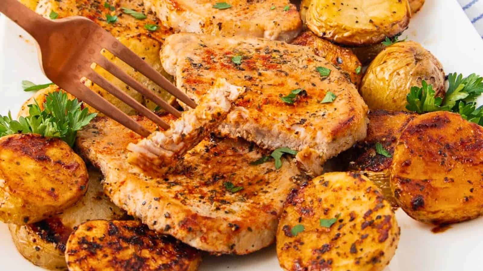 Seasoned pork chops and roasted potatoes garnished with parsley on a white plate, with a fork cutting a piece.