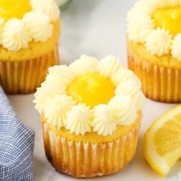 Three lemon cupcakes with white frosting and lemon filling, next to lemon slices and a blue checked napkin—perfect for brightening up your Easter recipes.