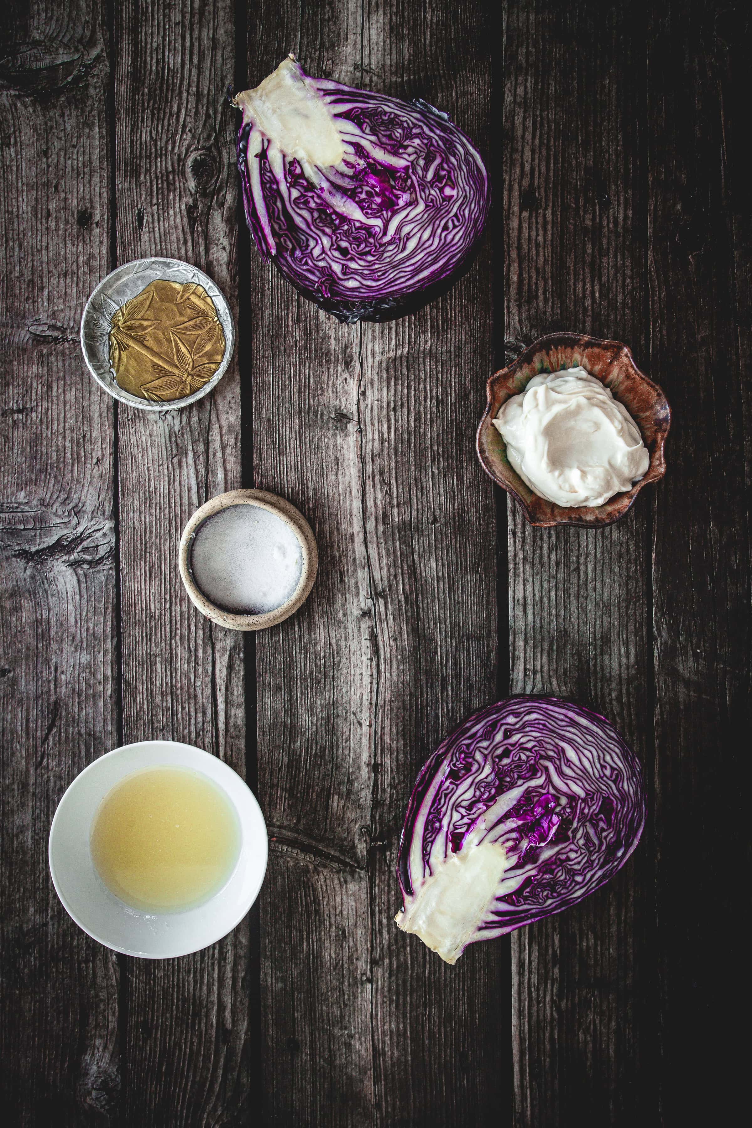 Halved red cabbage and small bowls of mayo, vinegar, salt, and mustard sit on a rustic wooden surface&mdash;ideal ingredients for a vibrant Mayo Purple Cabbage Slaw.