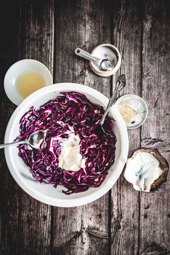 A bowl of Mayo Purple Cabbage Slaw sits on a rustic wooden table, surrounded by small bowls of ingredients, showcasing shredded red cabbage mixed with creamy mayonnaise.