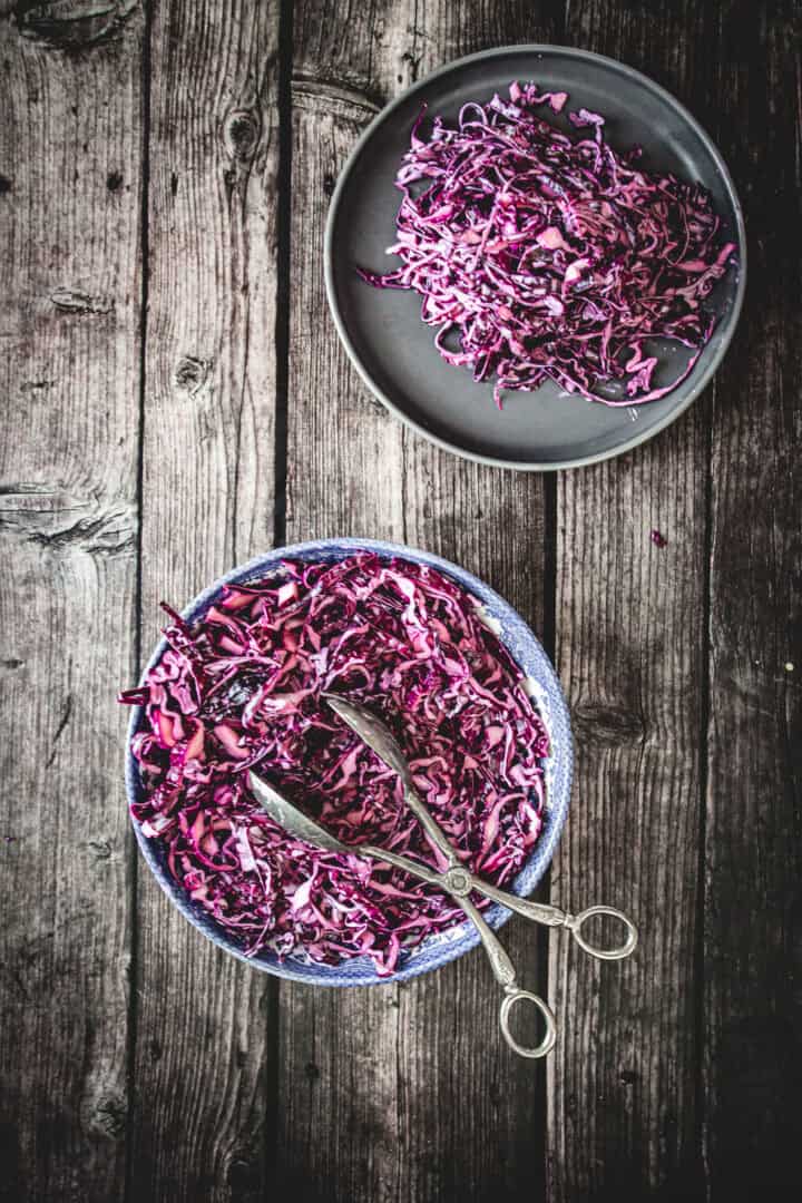 Shredded red cabbage on two plates with a pair of scissors, ready to be tossed into a creamy Mayo Purple Cabbage Slaw, all set on a rustic wooden table.