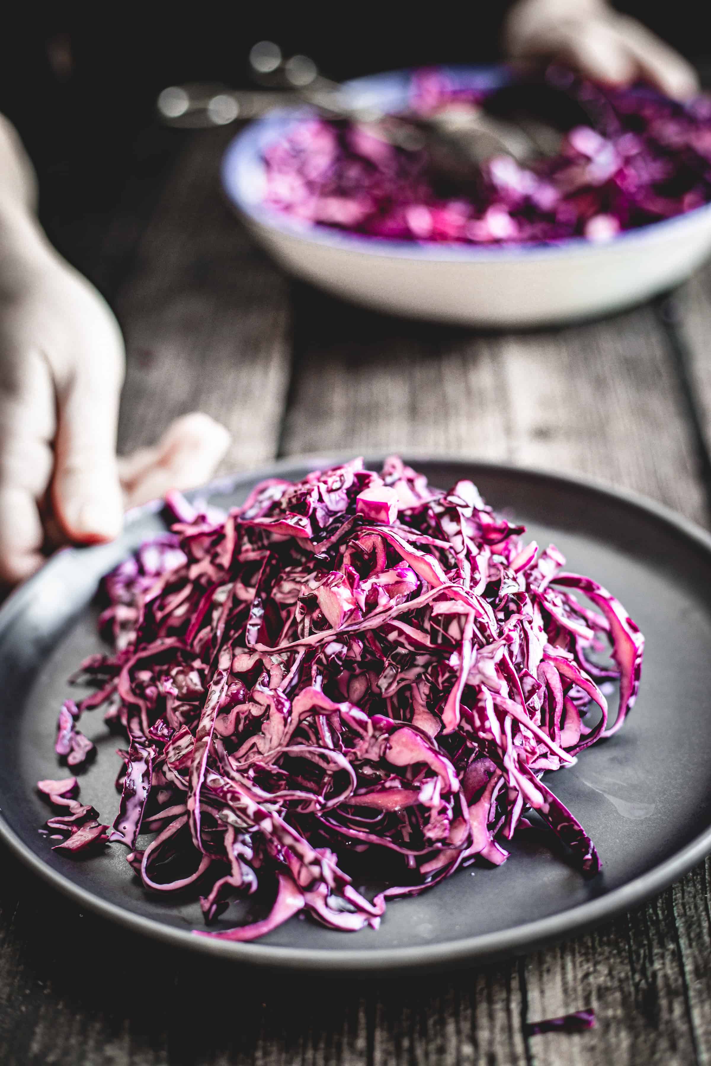 Shredded red cabbage on a dark plate, with a bowl of Mayo Purple Cabbage Slaw and hands in the background.