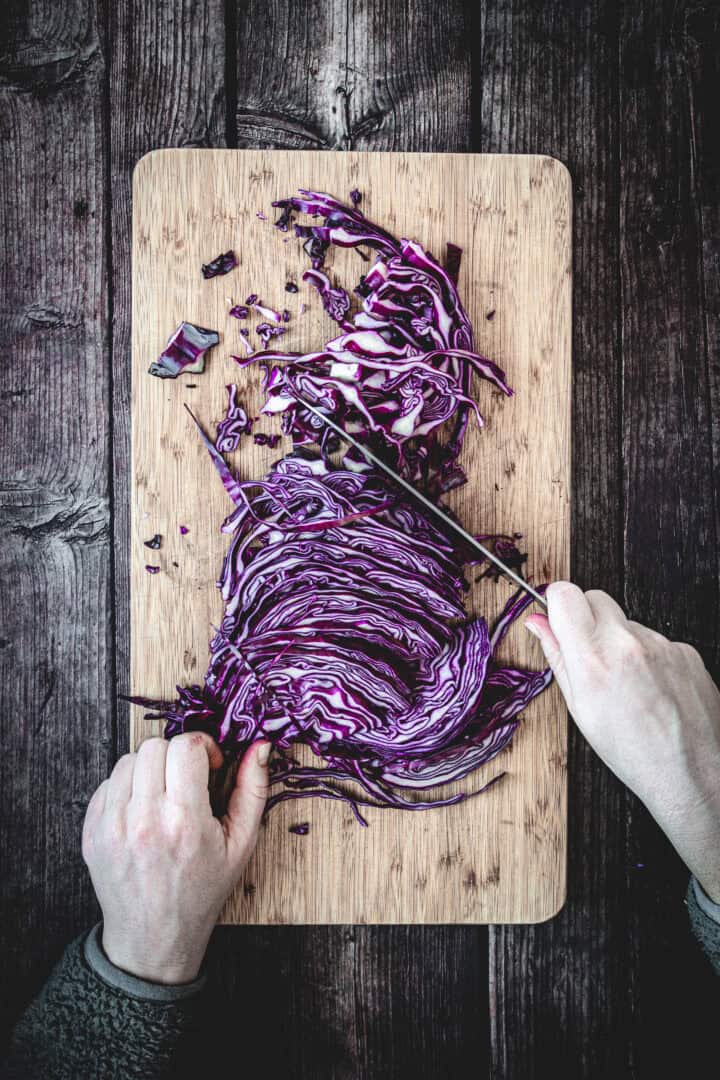 Top view of hands slicing purple cabbage on a wooden cutting board atop a dark wooden table, perfect for preparing fresh mayo purple cabbage slaw.