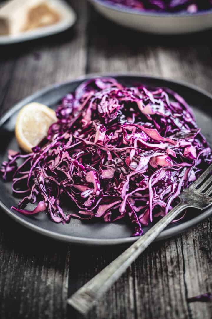 A plate of Mayo Purple Cabbage Slaw with a lemon wedge and a fork sits on a dark wooden table.