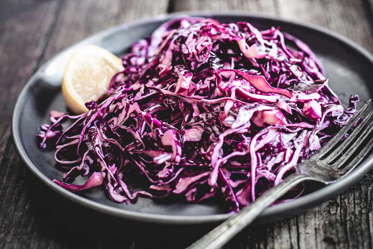 Mayo Purple Cabbage Slaw served as a shredded red cabbage salad with a lemon wedge on a dark plate, next to a fork, on a wooden table.