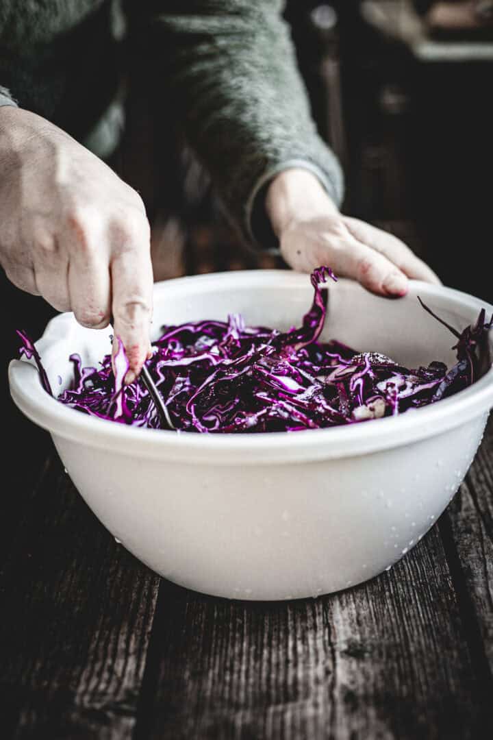 Hands mixing shredded purple cabbage for a Mayo Purple Cabbage Slaw in a large white bowl on a rustic wooden table.