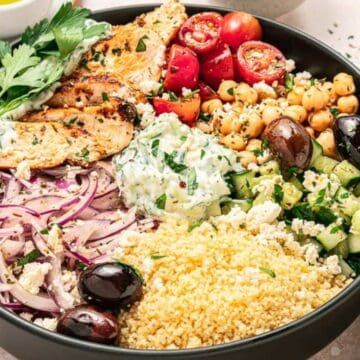 Bowl with grilled chicken, cherry tomatoes, chickpeas, couscous, cucumber, olives, onions, and tzatziki sauce.