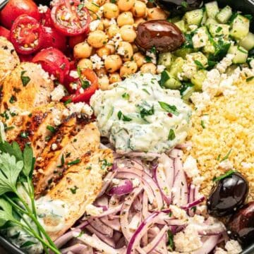 A colorful Mediterranean bowl with grilled chicken, couscous, tomatoes, cucumber, chickpeas, olives, and tzatziki.