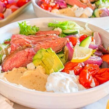 A bowl with sliced steak, avocado, tomatoes, hummus, tzatziki, greens, and vegetables.