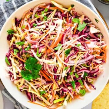 Bowl of colorful coleslaw with shredded cabbage, carrots, and cilantro, next to lime wedges and tortillas.
