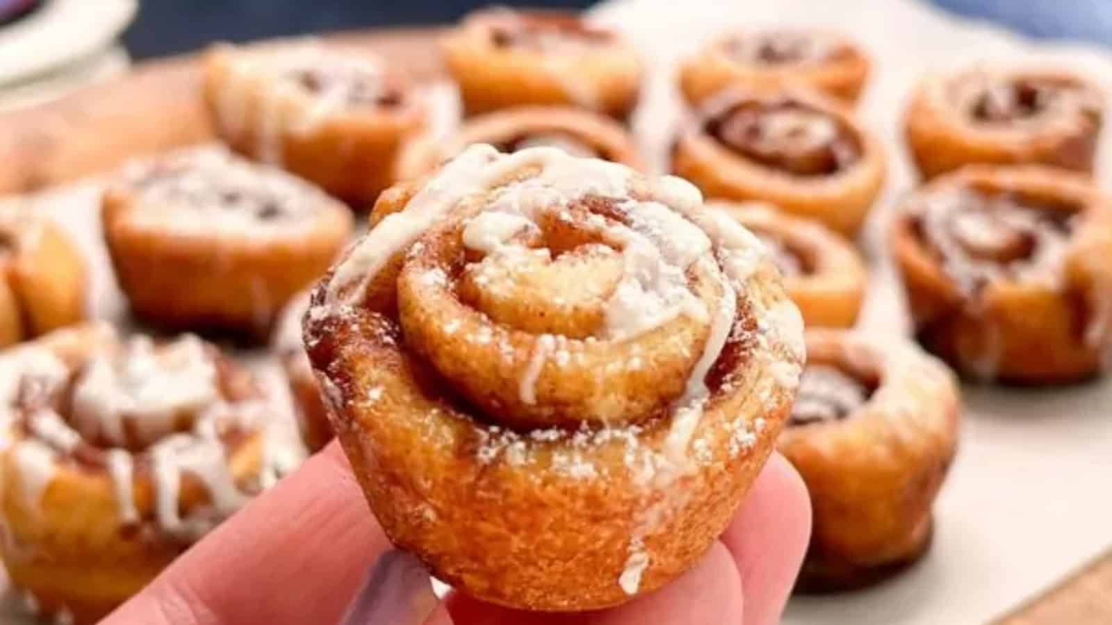 A hand holds a small, iced cinnamon roll, perfect as sweet appetizers, with more mini cinnamon rolls arranged on a tray in the background.