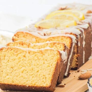 A loaf of lemon pound cake with icing, sliced on a wooden board, with lemons and almonds beside it.