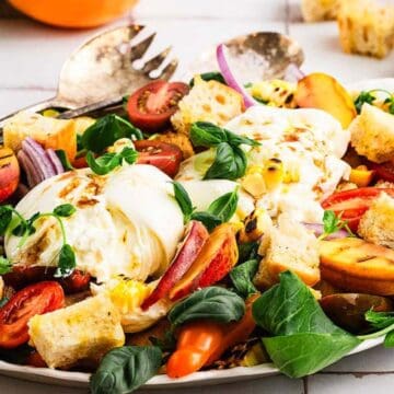 Colorful salad with mozzarella, tomatoes, peaches, croutons, greens, and red onions on a white platter.