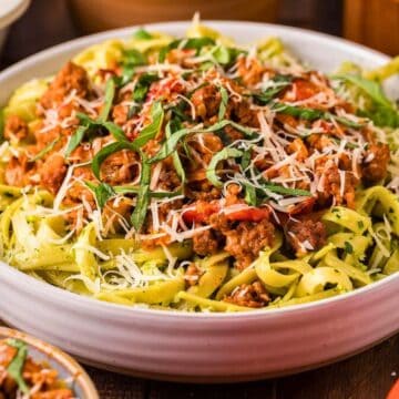 A bowl of fettuccine pasta topped with meat sauce, shredded cheese, and fresh basil leaves.