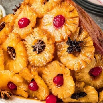 A baked ham garnished with pineapple rings, cherries, and star anise on a white plate.