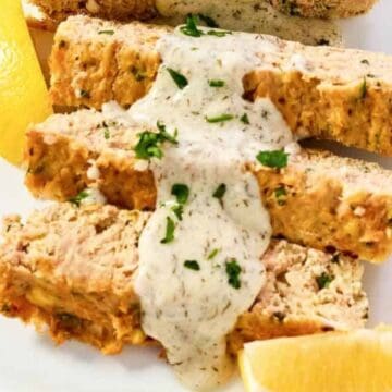 Slices of salmon loaf topped with creamy sauce and parsley, served with lemon wedges on a white plate.